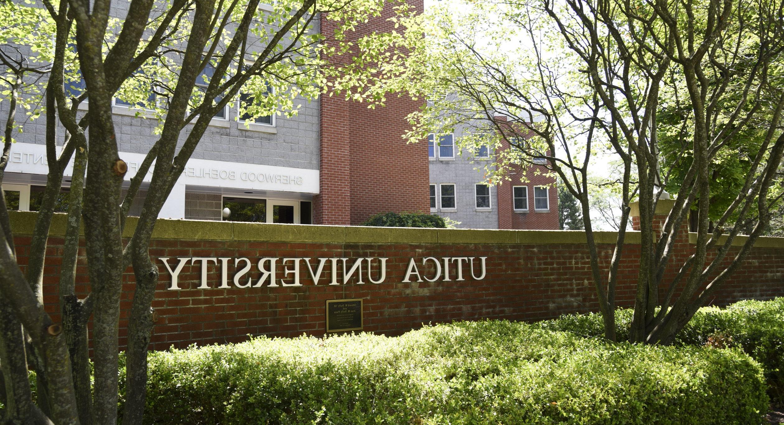 The words Utica University against a brick wall in front of a residence hall.