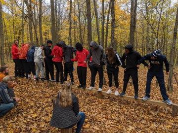 Students in the woods stand on a log holding hands.