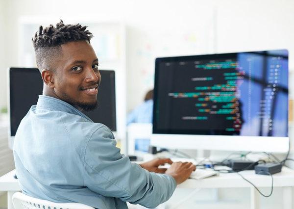 Cybersecurity student sits at computer, turning and smiling for camera.