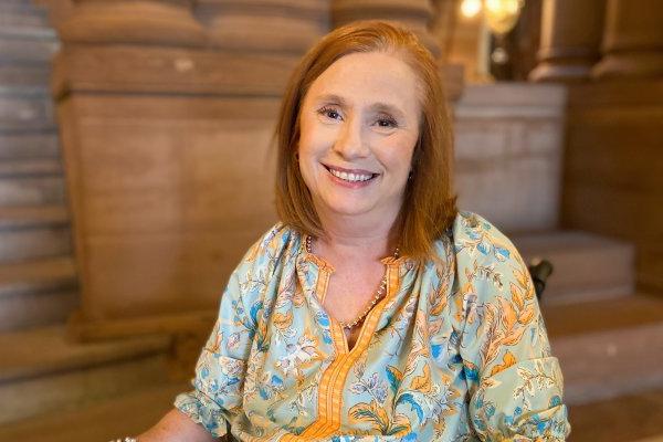 金伯利山, with red hair and a blue-yellow shirt, sits in her wheelchair inside the capital building in Albany, 微笑.